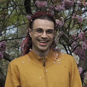 click for larger a person (troy) with pink hair and glasses smiles as pink petals fall from the cherry tree above across their face and onto their mustard-coloured cardigan